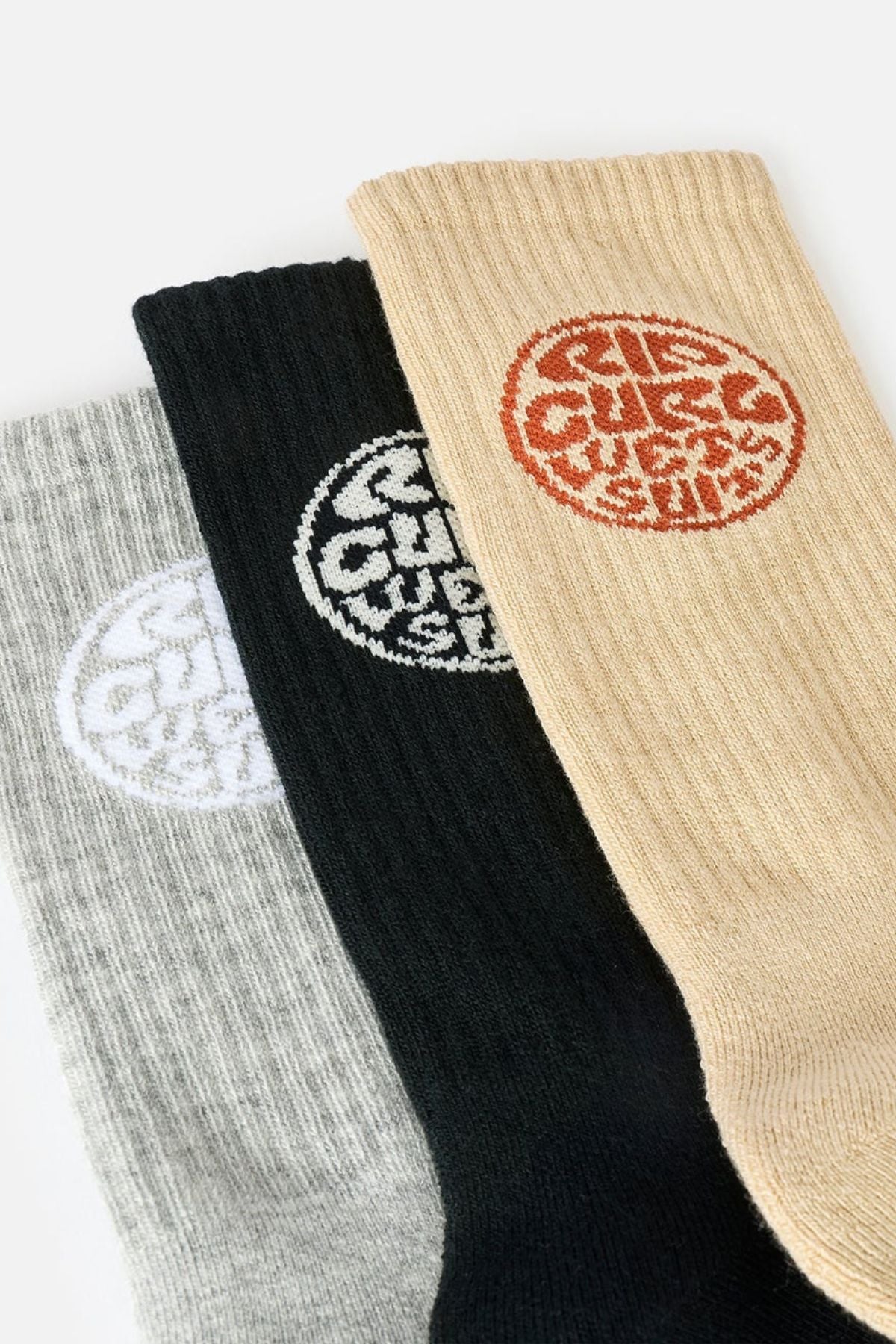 The Rip Curl Wetty Crew Sock 3 Pack, featuring three ribbed socks in gray, black, and beige—each with a circular logo near the top in white, black, and red respectively—are arranged side by side on a light background. These socks are designed for comfort with moisture-wicking fabric and built-in arch support.