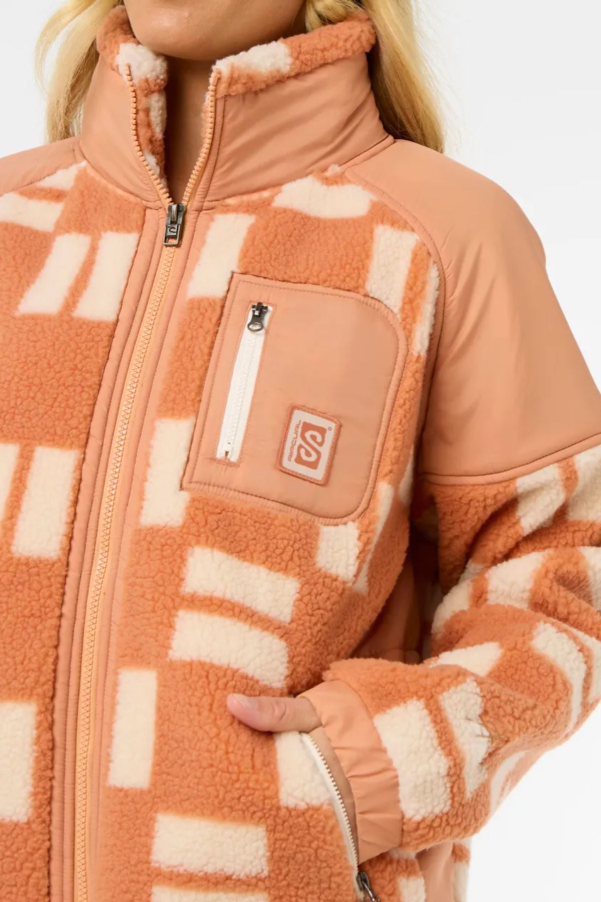 A woman wears the Rip Curl Anti-Series Shell Cove Fleece, featuring a relaxed fit, peach and cream geometric pattern, high collar, front zipper, zippered chest pocket, and side pockets. Only her lower face is visible.