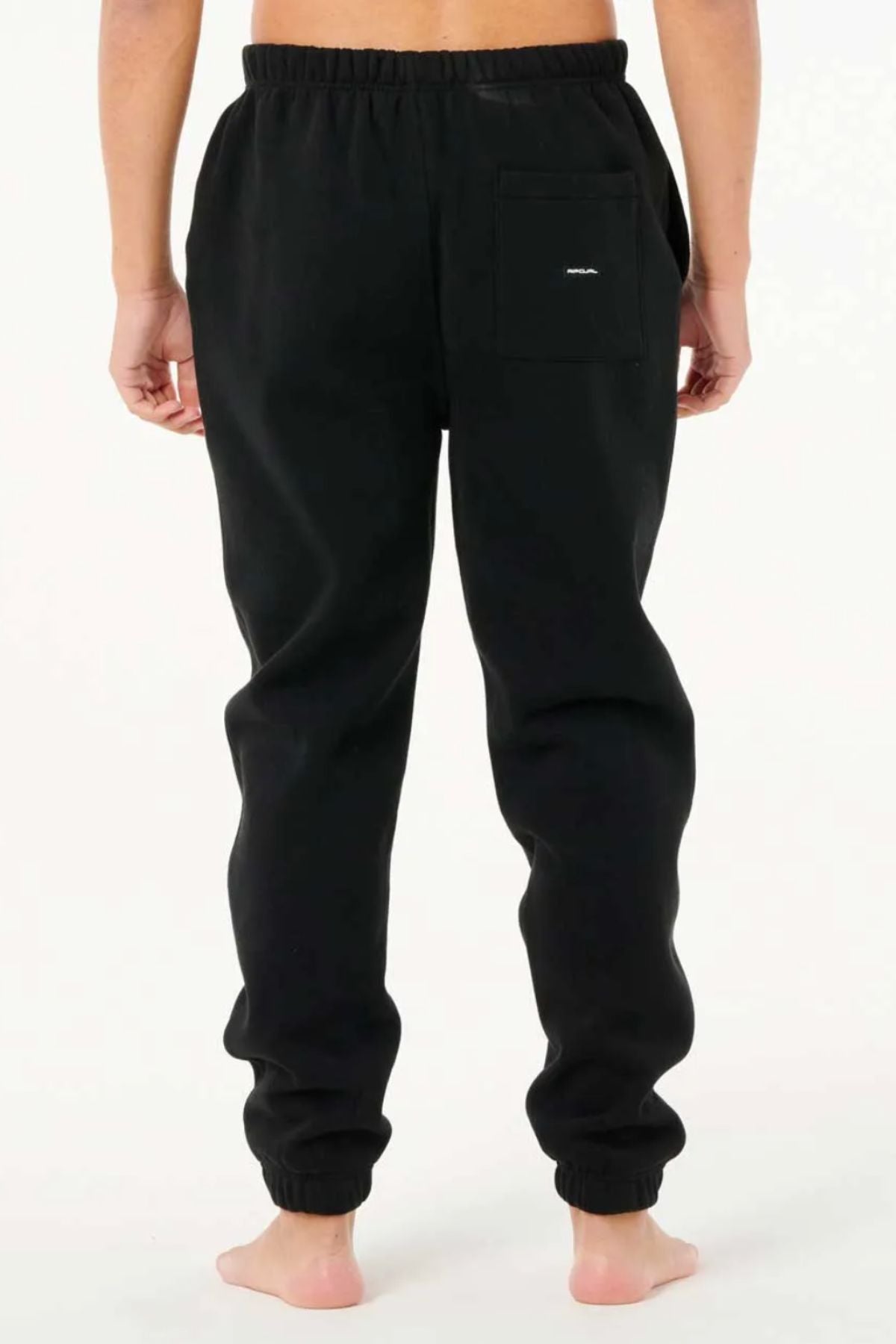A person stands barefoot on a light background, shown from the back, wearing black Rip Curl Dosed Up Trackpant joggers with an elastic waistband, elastic cuffs, a back pocket, and streetwear style.