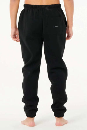 A person stands barefoot on a light background, shown from the back, wearing black Rip Curl Dosed Up Trackpant joggers with an elastic waistband, elastic cuffs, a back pocket, and streetwear style.