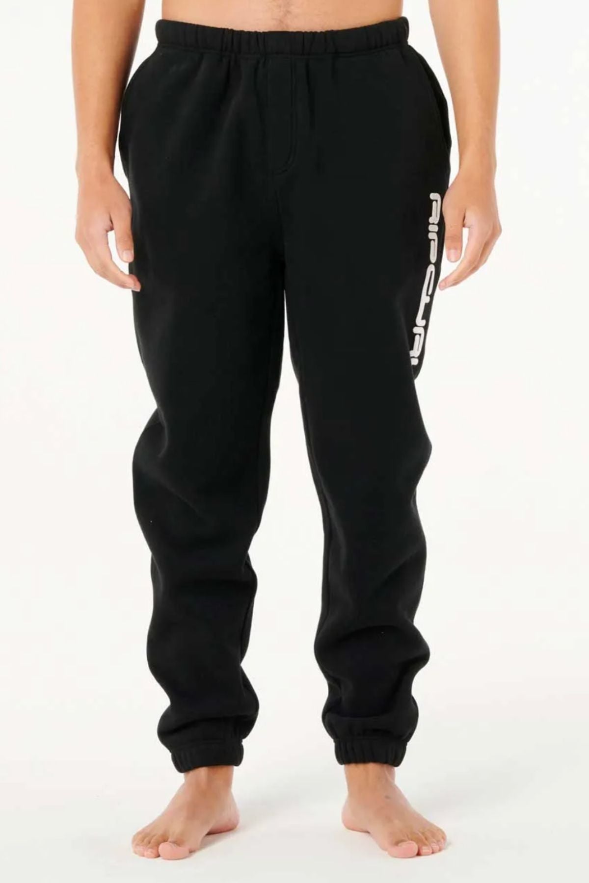 A person stands barefoot wearing black Rip Curl Dosed Up Trackpant joggers with white text on the left thigh, adding streetwear flair against a plain light background. The upper body and head are not visible.