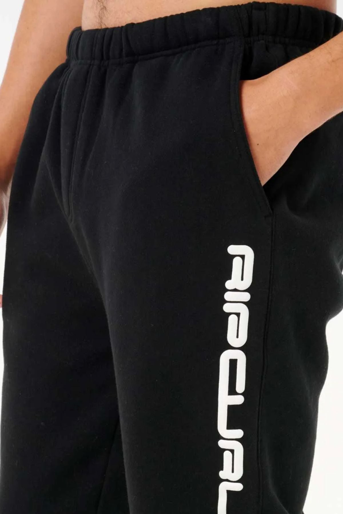 A person wears Rip Curl Dosed Up Trackpant black sweatpants with one hand in the pocket; made from a cotton-poly blend, these pants display bold white vertical text along the thigh for a trendy streetwear look.