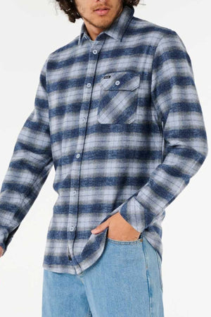 A person in the blue and gray plaid Rip Curl Grinners Flannel Shirt (100% cotton, relaxed fit, chest pocket) paired with light blue jeans stands with one hand in their pocket and their face partially out of frame.