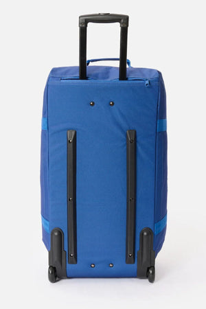 The Rip Curl Jupiter 80L Mixed Travel Bag is a blue, rectangular travel bag made from recycled polyester, featuring two black rails, wheels for upright standing, and a black telescoping handle. It is displayed against a plain white background.