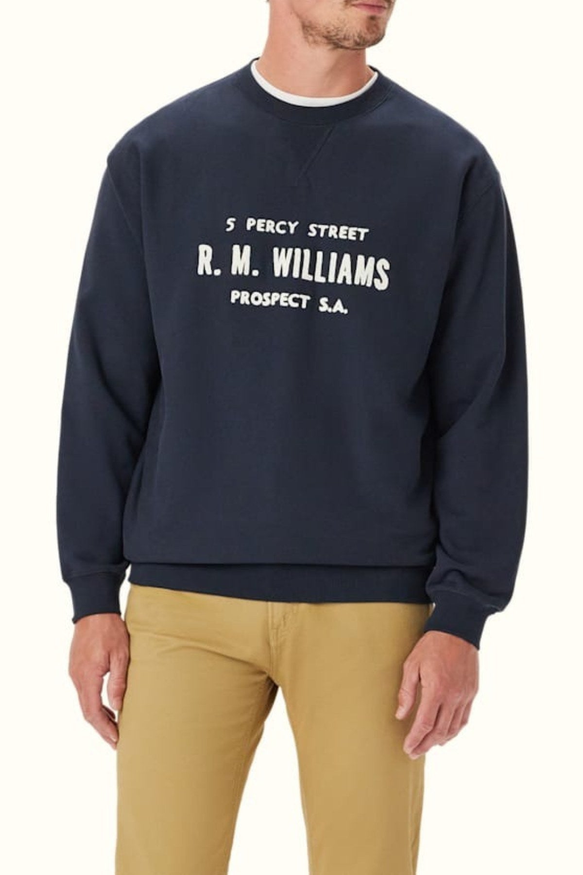 A man wears the RM Williams Bale Loopback Sweatshirt in relaxed-fit navy with white "5 Percy Street R.M. Williams Prospect S.A." embroidery, paired with beige pants. His face is partially out of frame.