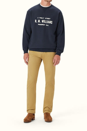 A person wears the RM Williams Bale Loopback Sweatshirt, a relaxed-fit navy blue organic cotton top with white "5 Percy Street R.M. Williams Prospect S.A." text, tan pants, and brown suede shoes against a plain light background.