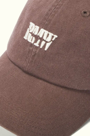 A close-up of the RM Williams Branded Twill Cap in brown organic cotton, featuring clear white embroidered text (upside down and distorted), visible stitching, textured fabric, and an adjustable strap.