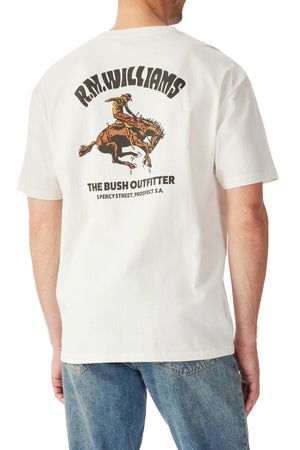 A person wears the RM Williams Cloudbuster T-Shirt, a white organic cotton jersey featuring a cowboy riding a bucking horse, inspired by Australian rodeos, with "R.M. Williams, The Bush Outfitter, Percy Street, Prospect S.A." text, paired with blue jeans.