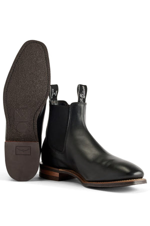 A pair of black RM Williams Comfort Craftsman Boots in pull-up leather with elastic side panels and pull tabs; one stands upright, the other lies on its side showing the textured sole and stacked heel—a classic, comfortable style.
