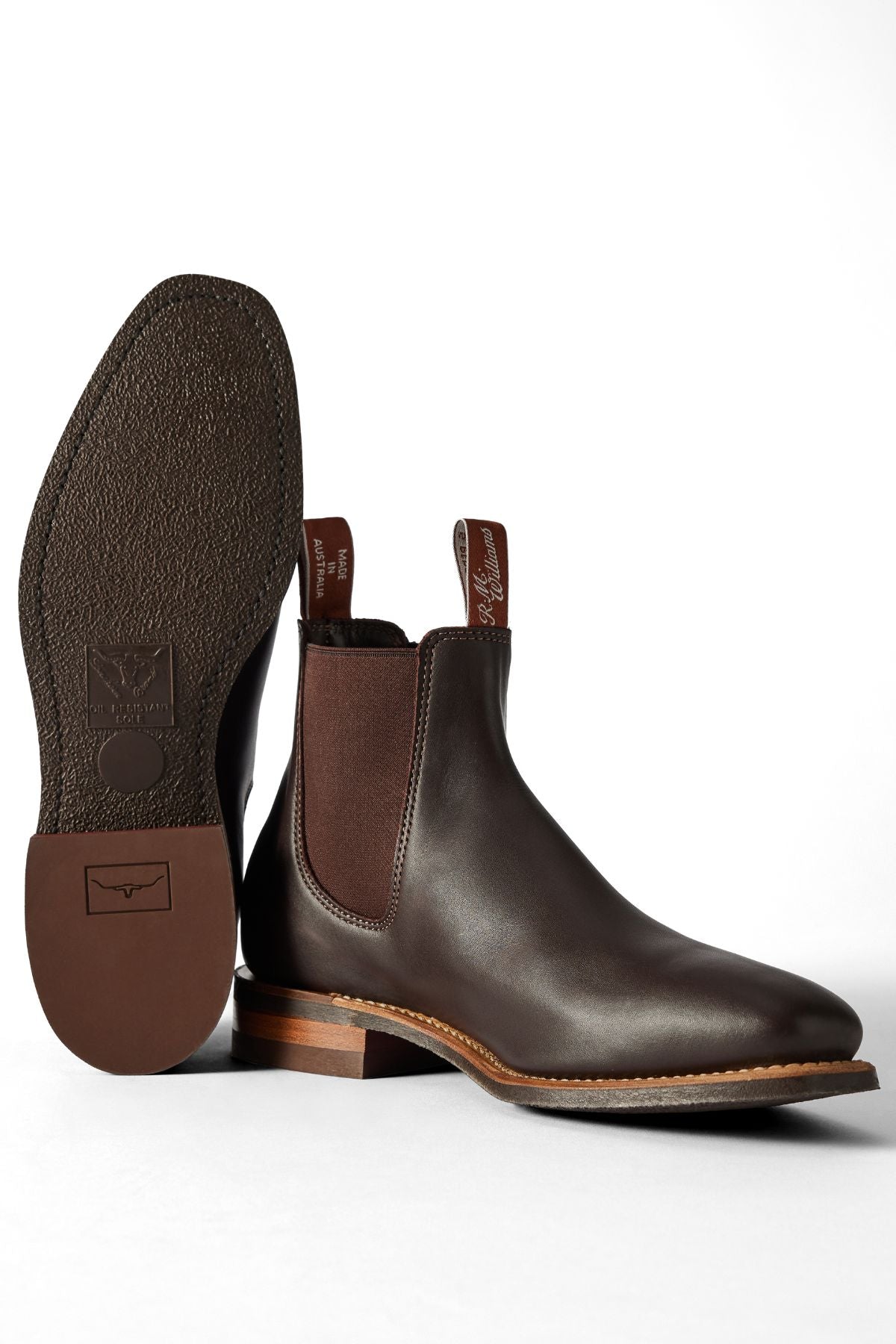 A pair of RM Williams Comfort Craftsman Boots in brown pull-up leather with elastic side panels. One boot stands upright as the other shows its textured rubber sole and wooden heel, set against a white background.