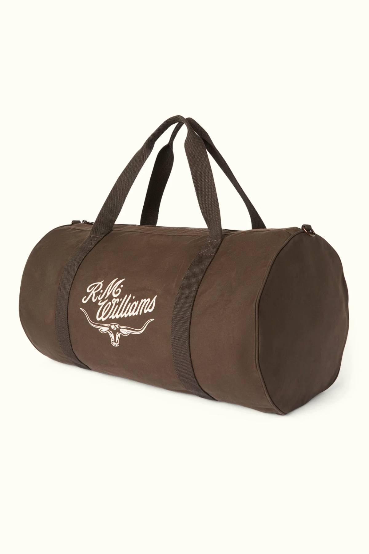 The RM Williams Ditty Bag, a brown cylindrical duffel made from durable waxed cotton canvas with two handles and the “R.M. Williams” logo in white script on the side, stands out against a plain light background.