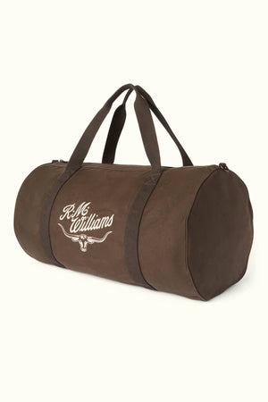 The RM Williams Ditty Bag, a brown cylindrical duffel made from durable waxed cotton canvas with two handles and the “R.M. Williams” logo in white script on the side, stands out against a plain light background.