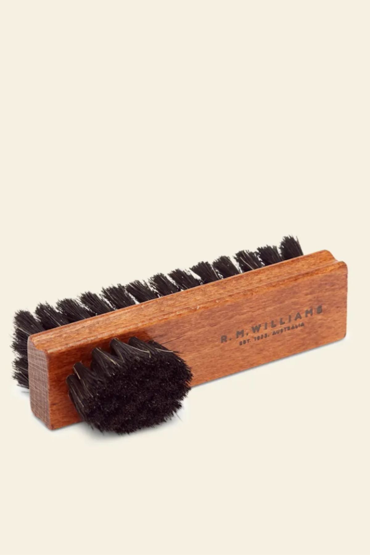 The RM Williams Double Sided Brush, featuring brown wood and black bristles, is displayed on a neutral background. One rectangular leather boot brush lies flat, with a smaller round brush angled in front of it.