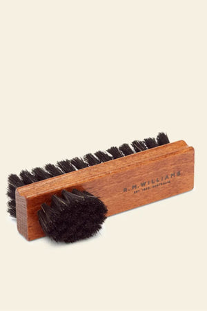 The RM Williams Double Sided Brush, featuring brown wood and black bristles, is displayed on a neutral background. One rectangular leather boot brush lies flat, with a smaller round brush angled in front of it.