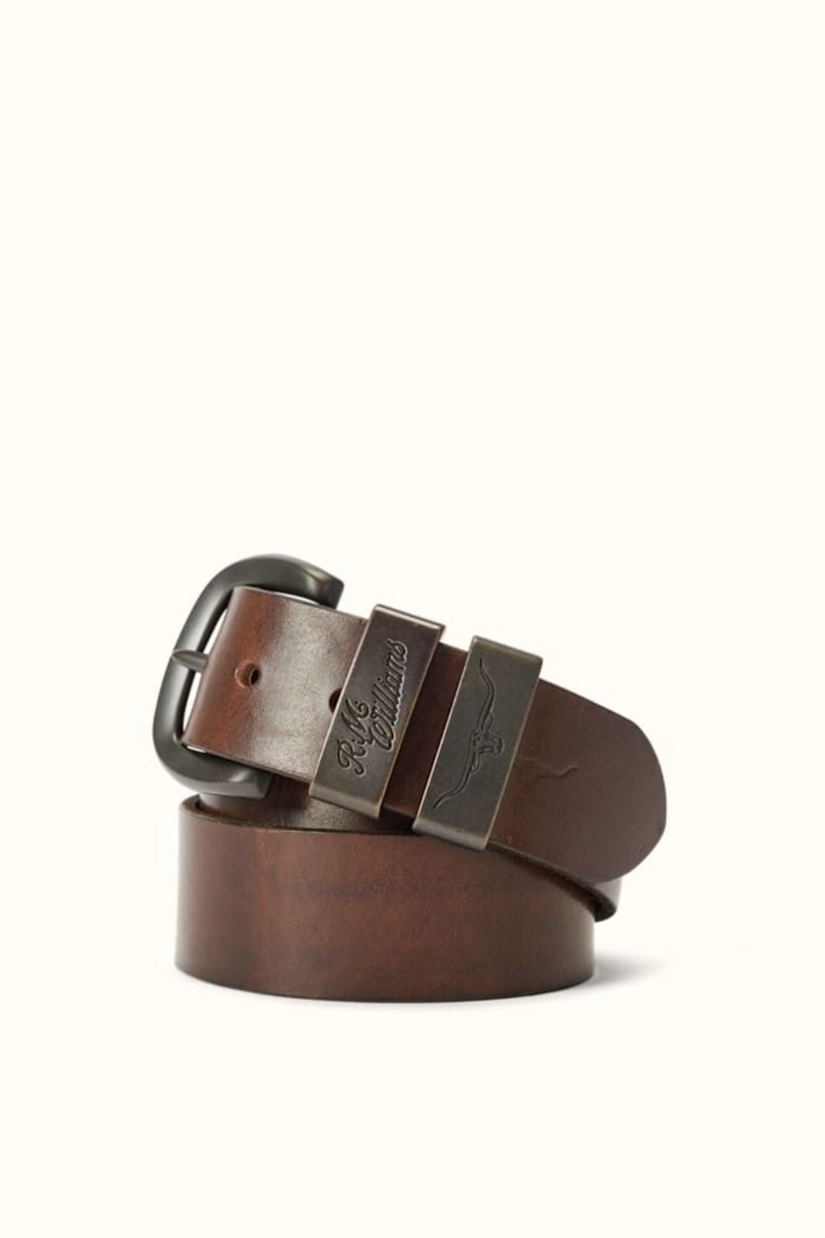 The RM Williams Drover Belt is a rolled leather belt with double keepers, a dark metal buckle, and two engraved metal clasps, shown against a plain light background.