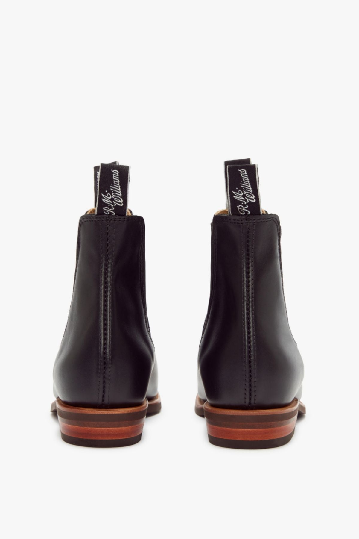 A pair of RM Williams Erica Boots in black full-grain leather, shown from the back with brown stacked heels and black pull tabs featuring white cursive text, displayed on a white background.