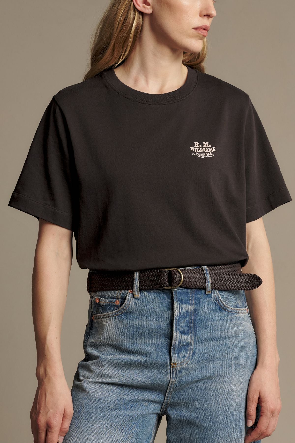 A person wearing an RM Williams Kandos Tee in black, tucked into blue jeans with a braided black belt, posing against a plain background.