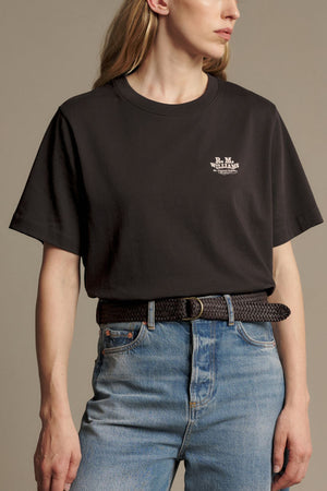 A person wearing an RM Williams Kandos Tee in black, tucked into blue jeans with a braided black belt, posing against a plain background.
