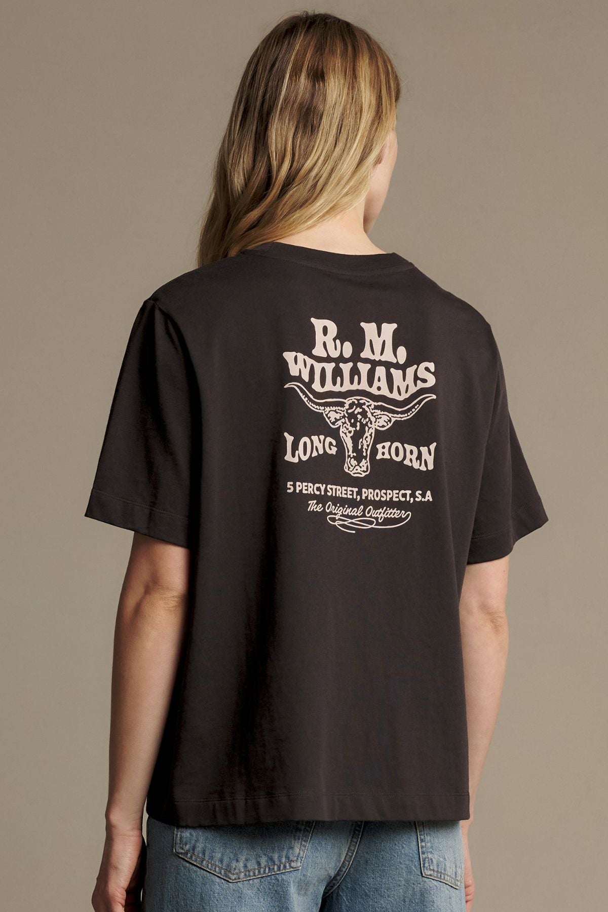 A person wearing the RM Williams Kandos Teeβa black, relaxed fit t-shirt featuring βR.M. Williams Long Horn,β a longhorn graphic, an address, and βThe Original Organiserβ text printed on the backβstanding against a plain background.