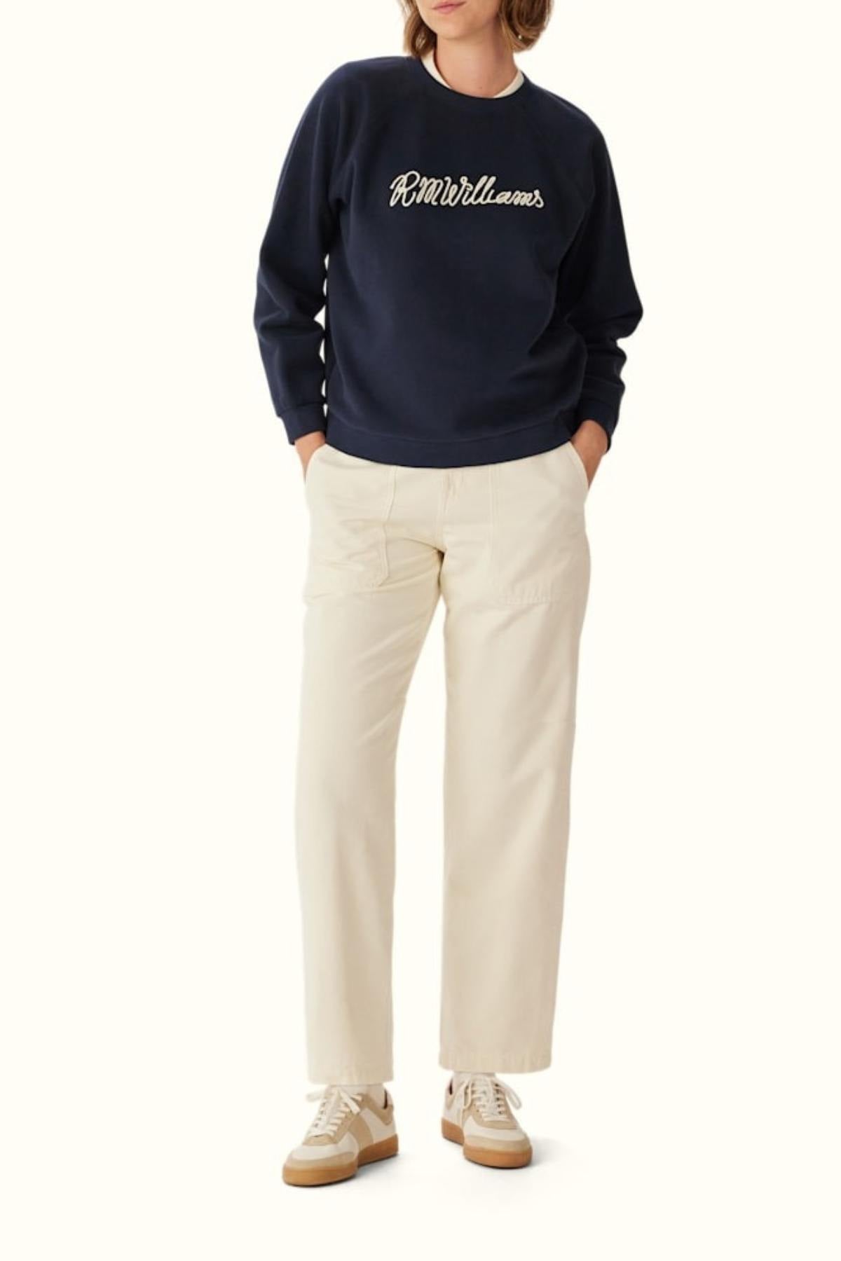 A person wears the RM Williams Lasso Brushed Sweatshirt in navy blue, paired with cream pants and beige sneakers. Their hands are in their pockets and their face is partially obscured.