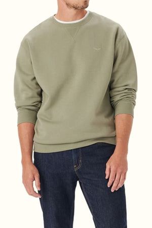 A man stands with his arms relaxed by his sides against a neutral background, wearing an RM Williams Mini Longhorn Sweatshirt in light olive green organic cotton and dark blue jeans.