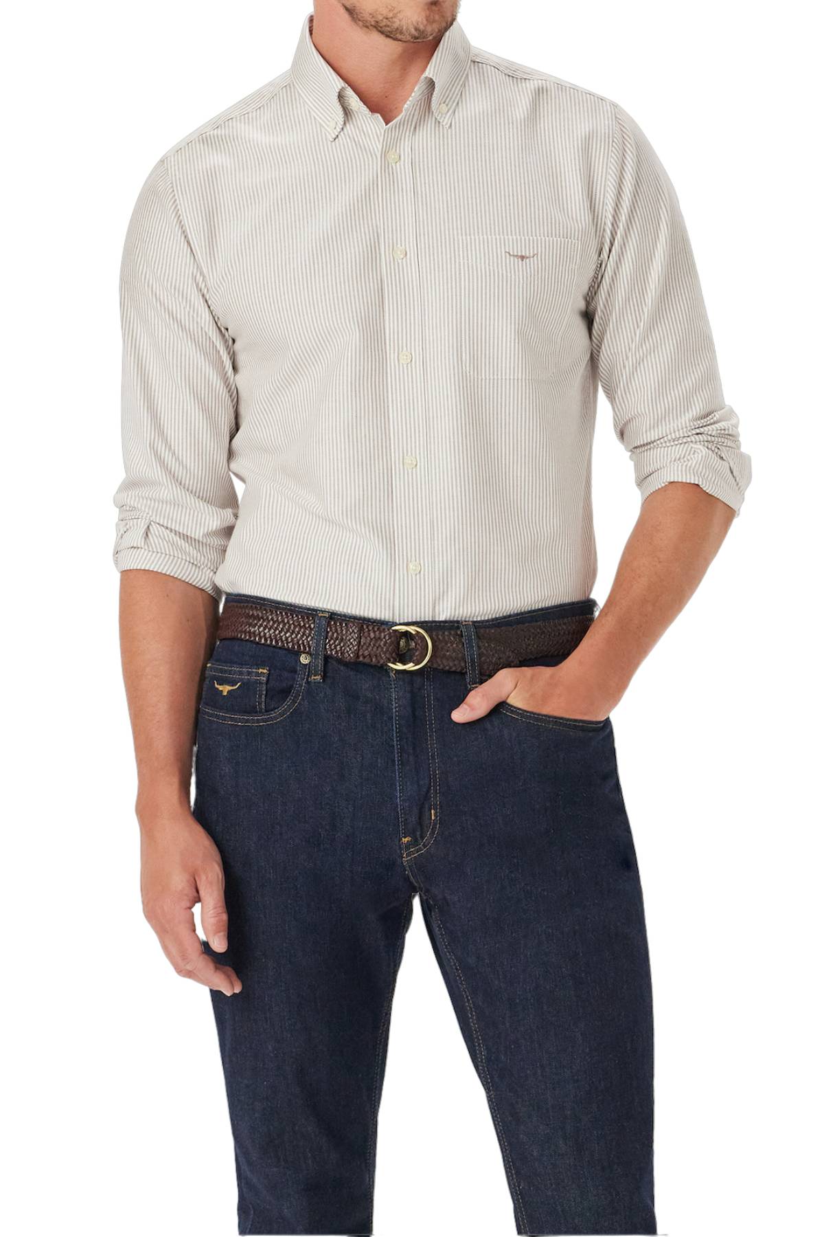 A man wears the RM Williams Regular Oxford Stripe Shirt—light-colored with thin vertical stripes in soft cotton—paired with dark blue jeans and a brown braided belt. His left hand is in his pocket, and his head is not visible.