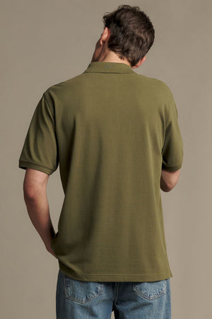 A person wearing the RM Williams Rod Polo in olive green Australian cotton, featuring a ribbed collar, and light blue jeans is standing with their back to the camera, head slightly turned to the left. The background is plain and neutral.