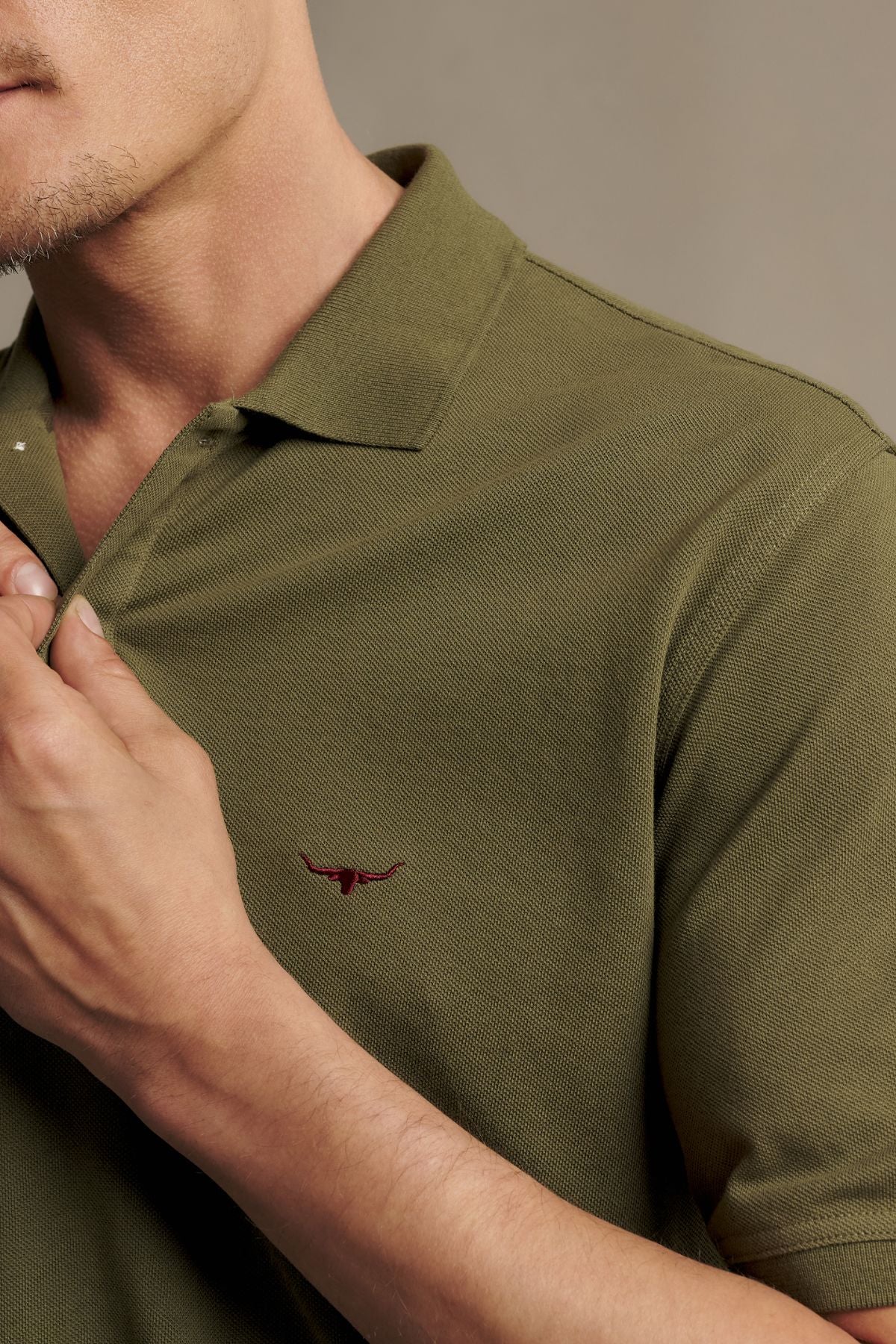 A man wearing the RM Williams Rod Polo in olive green—made from Australian cotton, featuring a small red bull head logo and subtle Longhorn embroidery on the chest—adjusts the ribbed collar with his hand.