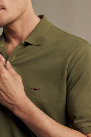 A man wearing the RM Williams Rod Polo in olive green—made from Australian cotton, featuring a small red bull head logo and subtle Longhorn embroidery on the chest—adjusts the ribbed collar with his hand.