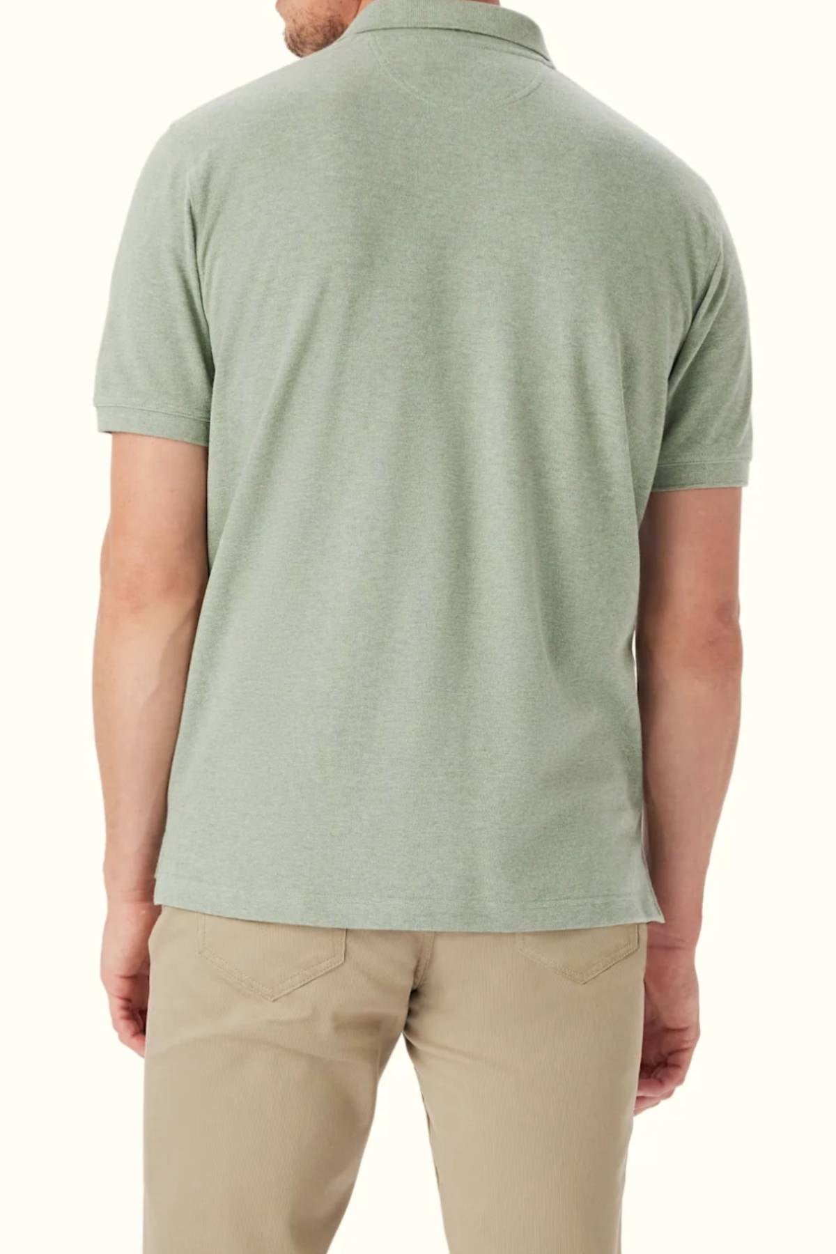 A man is seen from behind wearing the RM Williams Rod Polo in light green organic cotton with Longhorn embroidery, paired with beige pants. The background is plain and light-colored.