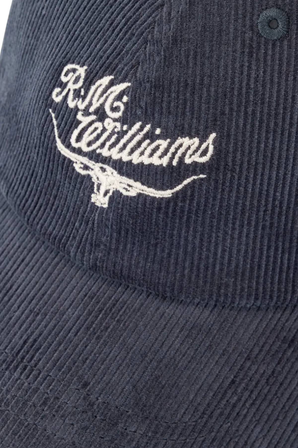 A close-up of the RM Williams Script Longhorn Cord Cap, a dark organic cotton corduroy hat featuring white "R.M. Williams" embroidery above a stylized longhorn logo.