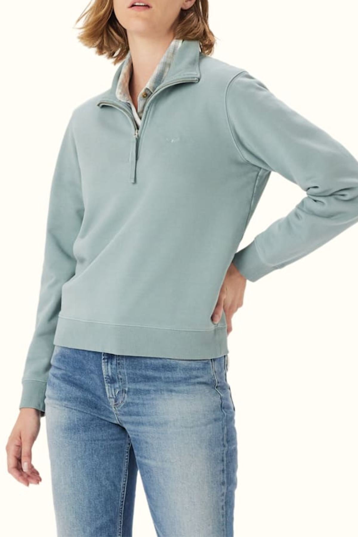 Wearing the RM Williams Trickett Zip Neck Sweatshirt in light blue with blue jeans, a person stands against a plain background, one hand on their hip and their face partially out of frame.