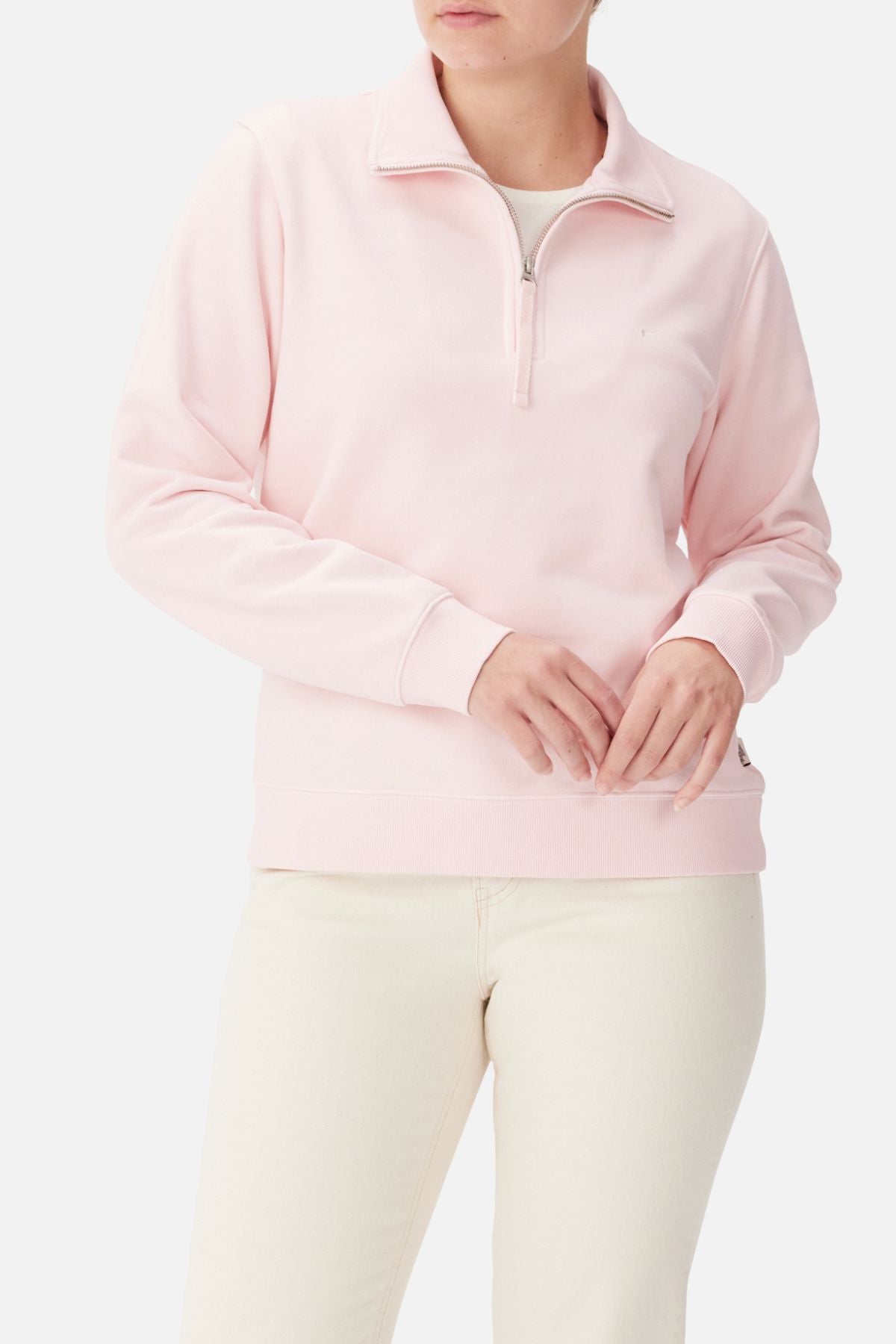 A person in the RM Williams Trickett Zip Neck Sweatshirt (light pink organic cotton) with off-white pants stands against a plain background, hands clasped at the waist, face partially out of frame.