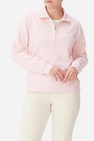 A person in the RM Williams Trickett Zip Neck Sweatshirt (light pink organic cotton) with off-white pants stands against a plain background, hands clasped at the waist, face partially out of frame.