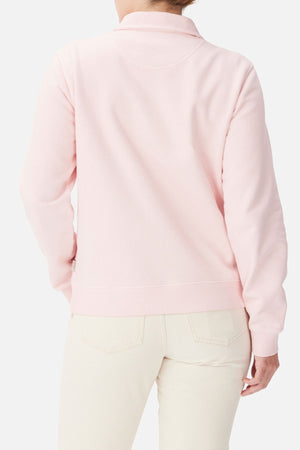 A person is seen from behind against a plain background, wearing the RM Williams Trickett Zip Neck Sweatshirt in light pink with off-white pants. Their left hand rests on their hip.