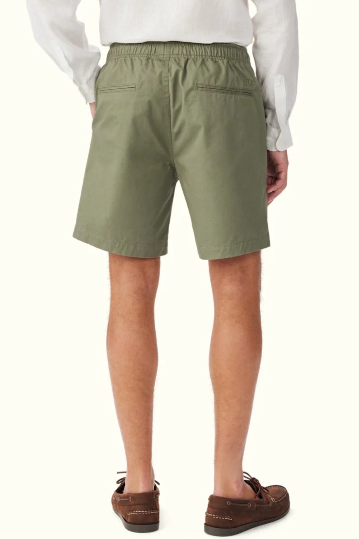 A person wearing RM Williams Washed Rugby Shorts in olive green, a white long-sleeve shirt, and brown boat shoes stands with their back to the camera against a plain light background.