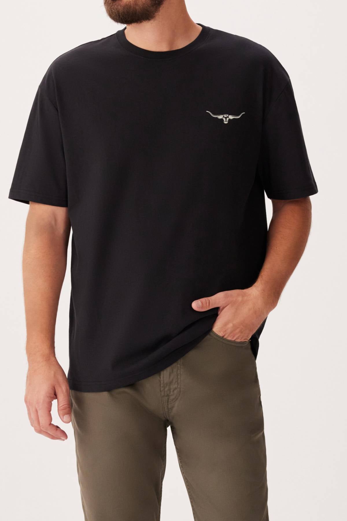 A man wearing the RM Williams 1932 T-Shirt, a black ribbed crew neck featuring a small white longhorn print on the left chest, paired with olive green pants and standing with one hand in his pocket.