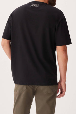 A person is shown from the back wearing an RM Williams 1932 T-Shirt in plain black organic cotton with a ribbed crew neck and a small white label near the neckline, paired with olive green pants against a plain light background.