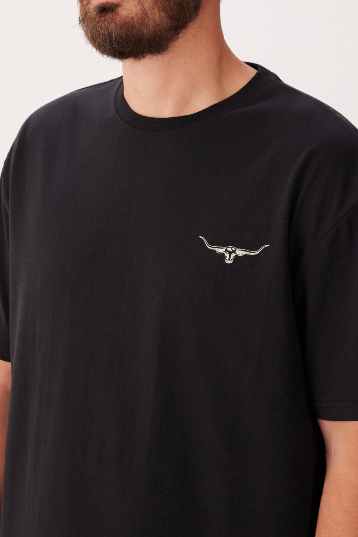 A person wearing the RM Williams 1932 T-Shirt—a black, ribbed crew neck with a small embroidered longhorn skull logo on the left chest. Only the lower half of their beard and jawline are visible; their face is partially out of frame.