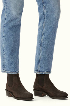 A person wearing light blue jeans and RM Williams Comfort Lady Yearling Boot in dark brown waxed suede with low heels, elastic side panels, and a Goodyear welt, standing on a neutral background.