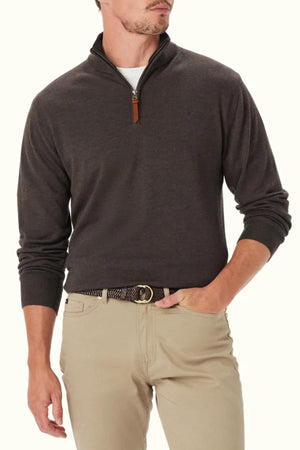 A man wearing the RM Williams Ernest 1/4 Zip Merino Knit in dark brown over a white shirt, paired with beige pants and a brown belt, stands with one hand in his pocket against a plain background.