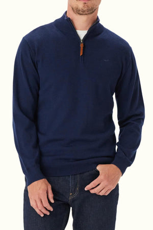 A man wears the RM Williams Ernest 1/4 Zip Merino Knit in navy blue with a brown zipper pull, paired with dark blue jeans, standing against a plain white background.