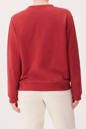 A person is seen from the back with arms relaxed, wearing the RM Williams Heritage Brushed Sweatshirt in plain red and light beige pants.