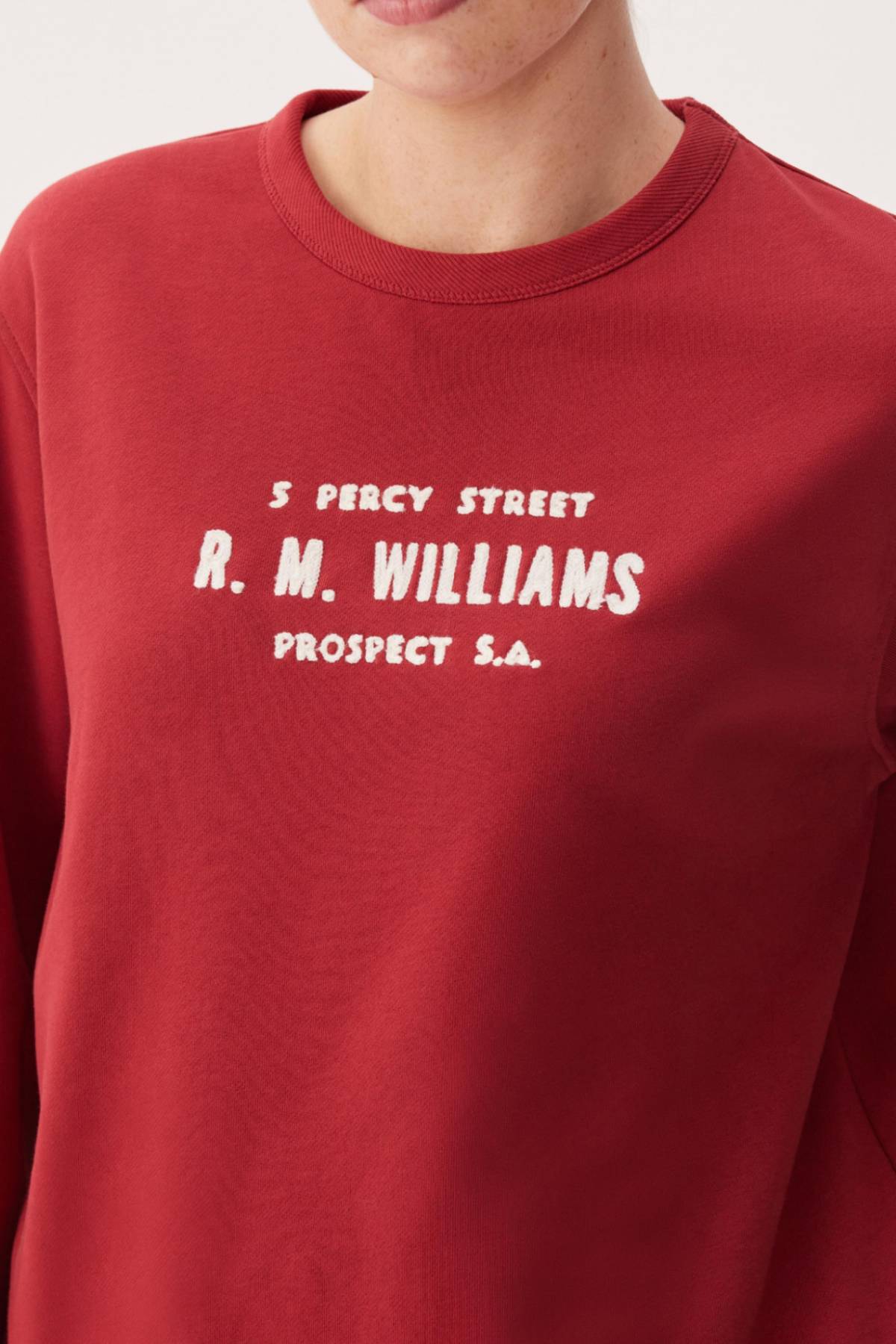 A person wears the RM Williams Heritage Brushed Sweatshirt in red, featuring white "5 Percy Street R. M. Williams Prospect S.A." embroidery. Only their lower face and upper torso are visible. Made from Australian Good Earth Cotton for comfort and style.