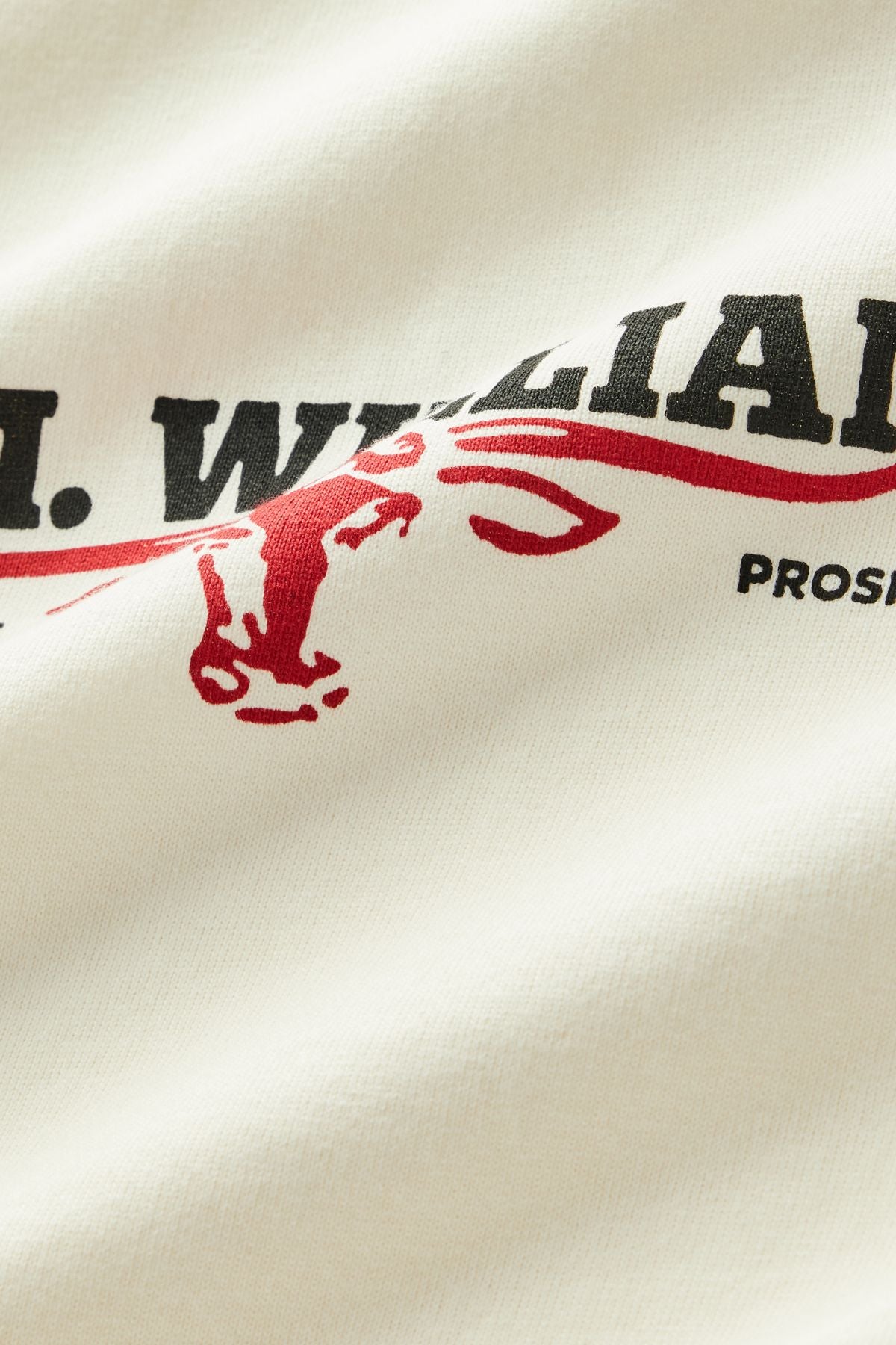 A close-up of the RM Williams Holt Tee’s white fabric, featuring bold black text and a red outline drawing of a donkey’s head. Some partially visible text reads “…WILLIAMS” and “PROS…”.