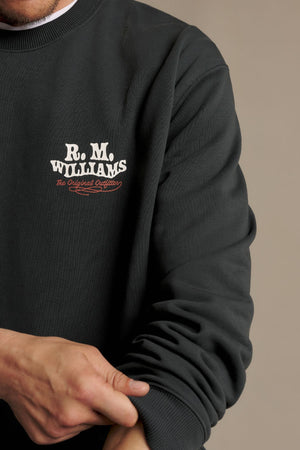 A person wearing the RM Williams Kandos Crew—a dark sweatshirt featuring “R.M. Williams The Original Outfitters” on the chest—rolls up their sleeve against a plain background.