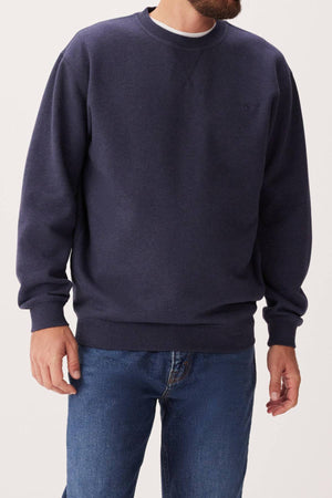 A man wears the RM Williams Mini Longhorn Sweatshirt, a navy blue ribbed crew neck made from an organic cotton blend, paired with blue jeans. He stands against a plain light background with his head partially out of frame.