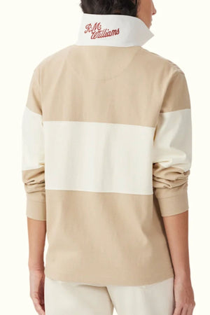 A person, facing away with rolled-up sleeves, wears the RM Williams Narran Panel Rugby—a beige and white heavyweight organic cotton jersey featuring a wide back stripe and a turned-up collar embroidered with the Heritage logo in red.