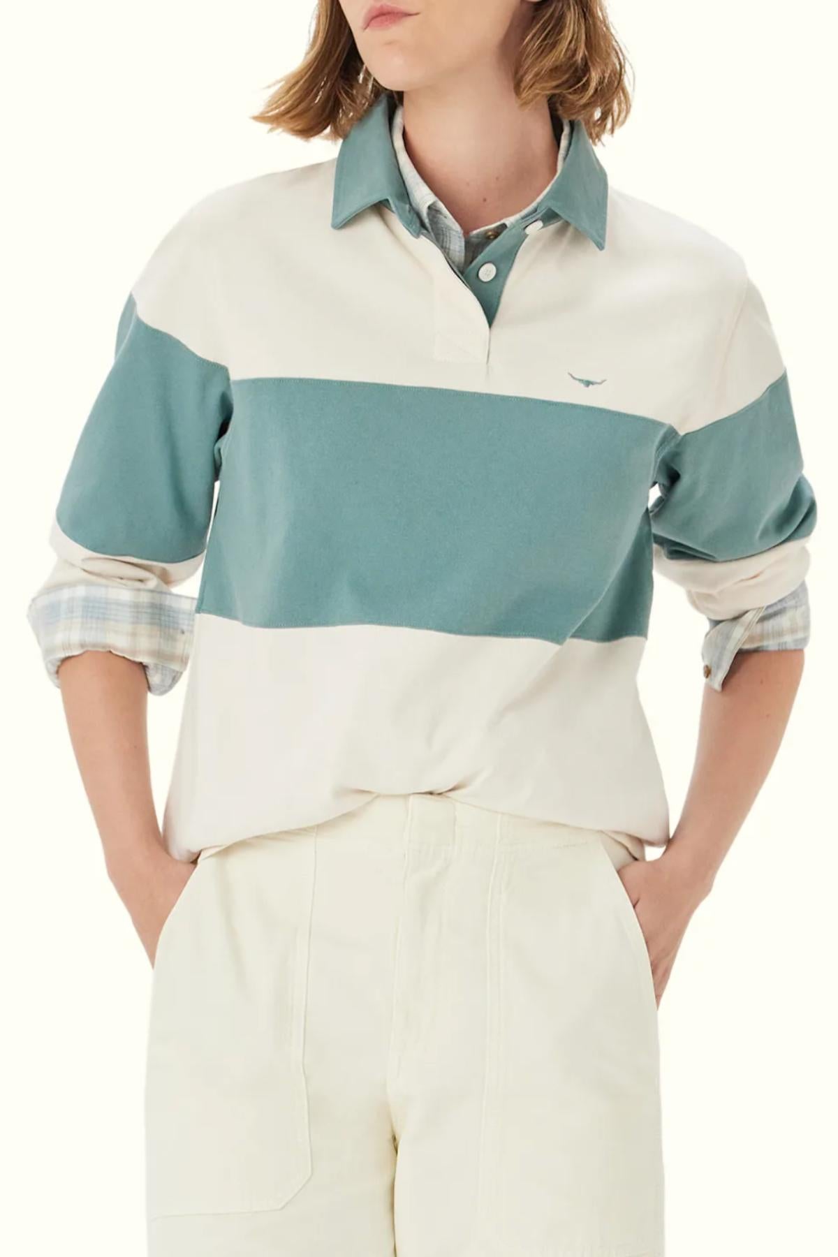 A person wearing the RM Williams Narran Panel Stripe Rugby, a green and white striped organic cotton rugby shirt, layered over a plaid collared shirt with hands in pockets, shown from shoulders to thighs.