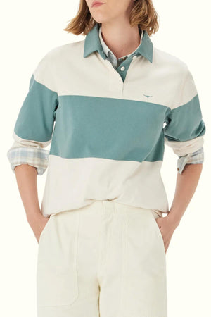 A person wearing the RM Williams Narran Panel Stripe Rugby, a green and white striped organic cotton rugby shirt, layered over a plaid collared shirt with hands in pockets, shown from shoulders to thighs.
